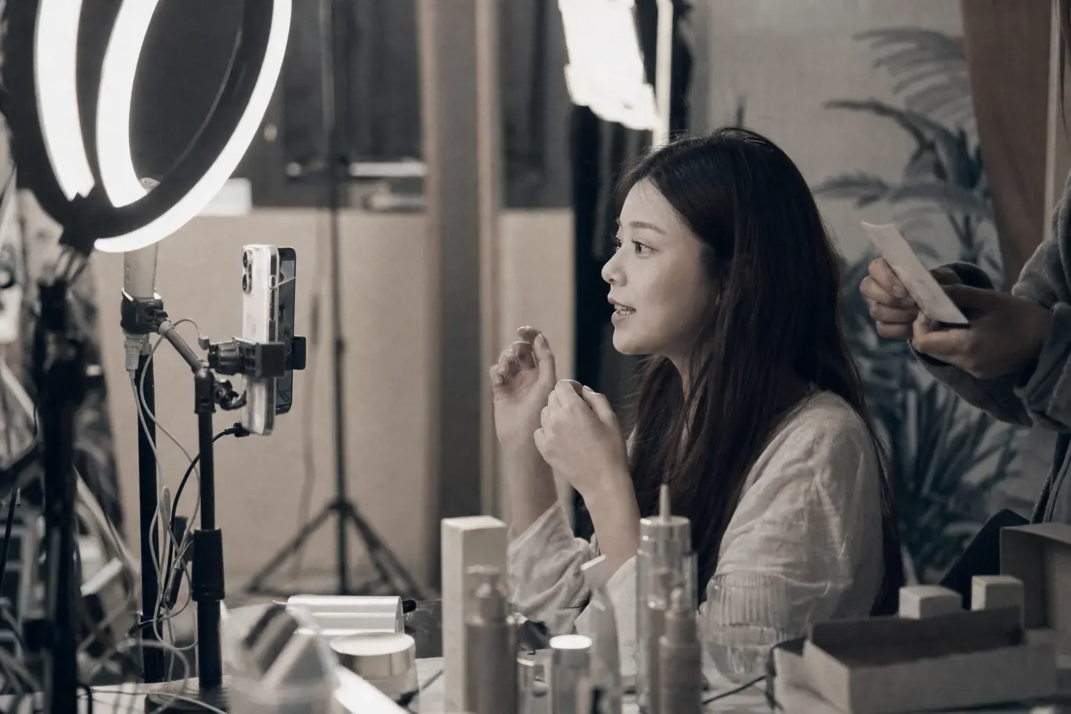A livestream host talking to a phone on a tripod under a ring light, product samples lined up in front of her