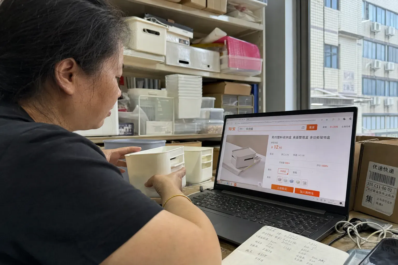 A Chinese merchant in a Yiwu showroom compares a Taobao product page with household storage samples beside her laptop.