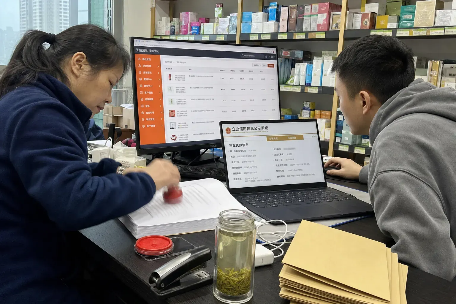 In a Chengdu distributor's office, a woman stamps a red company seal on a printed contract while a colleague checks the company's business license record on a laptop.