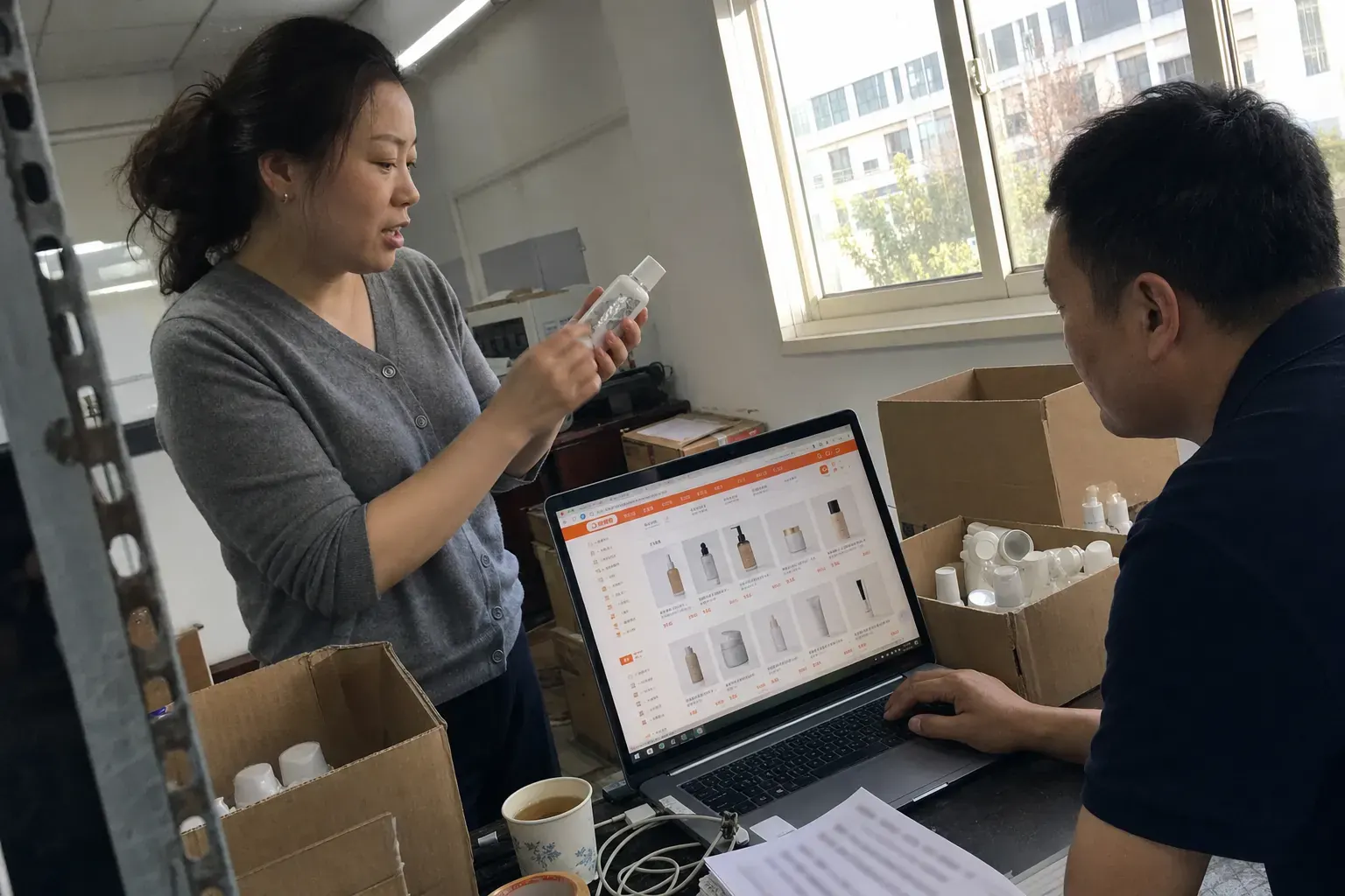 Two staff in a Shanghai Fengxian cosmetics stockroom compare a bottle with its code scratched off against an online shop listing on a laptop.