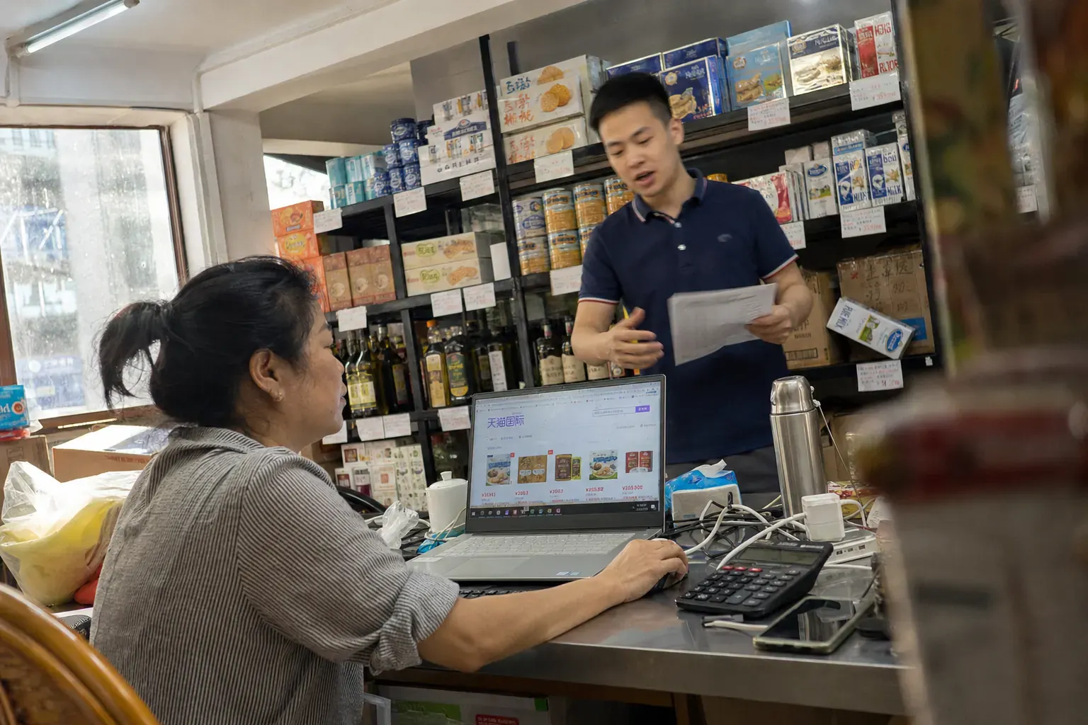 A woman at a cluttered desk in a Guangzhou food distributor's showroom checks a Tmall Global store page on a laptop while a colleague holds a printed price list