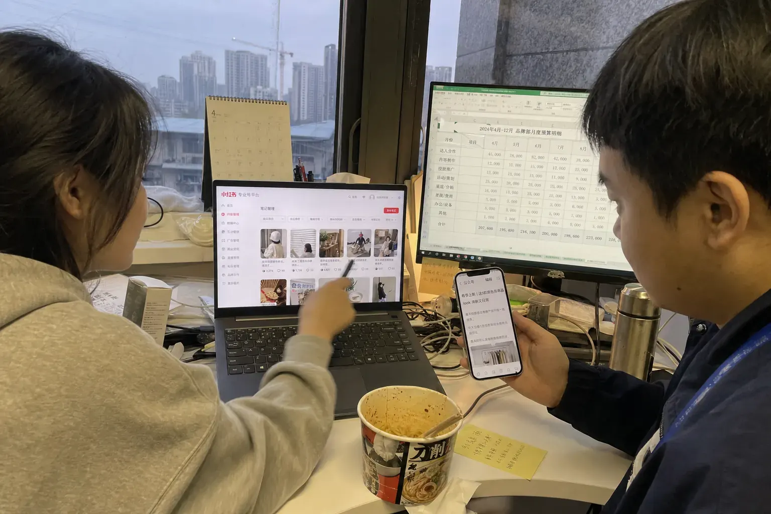 Two young staff at a cluttered desk in a Wuhan office compare Xiaohongshu brand notes on a laptop and a WeChat article draft on a phone, a budget spreadsheet on the monitor behind them