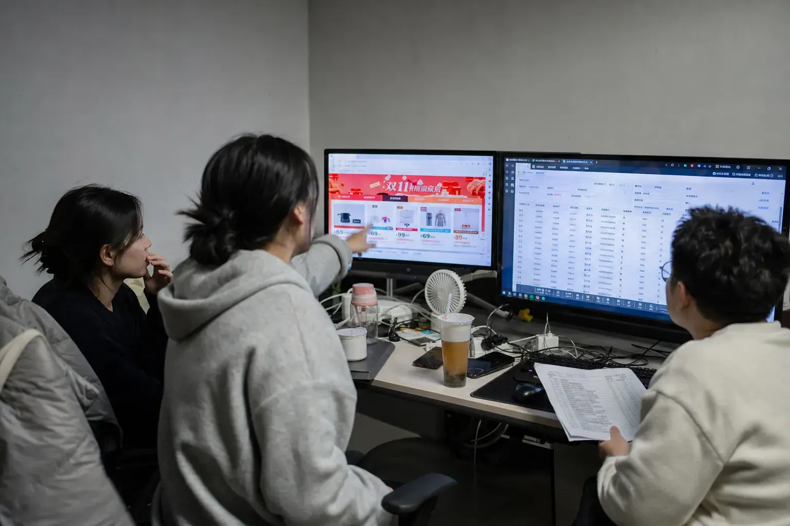 Three staff in a Hangzhou cross-border ecommerce office check a Chinese Tmall Global storefront and a warehouse dashboard on two cluttered monitors late in the evening