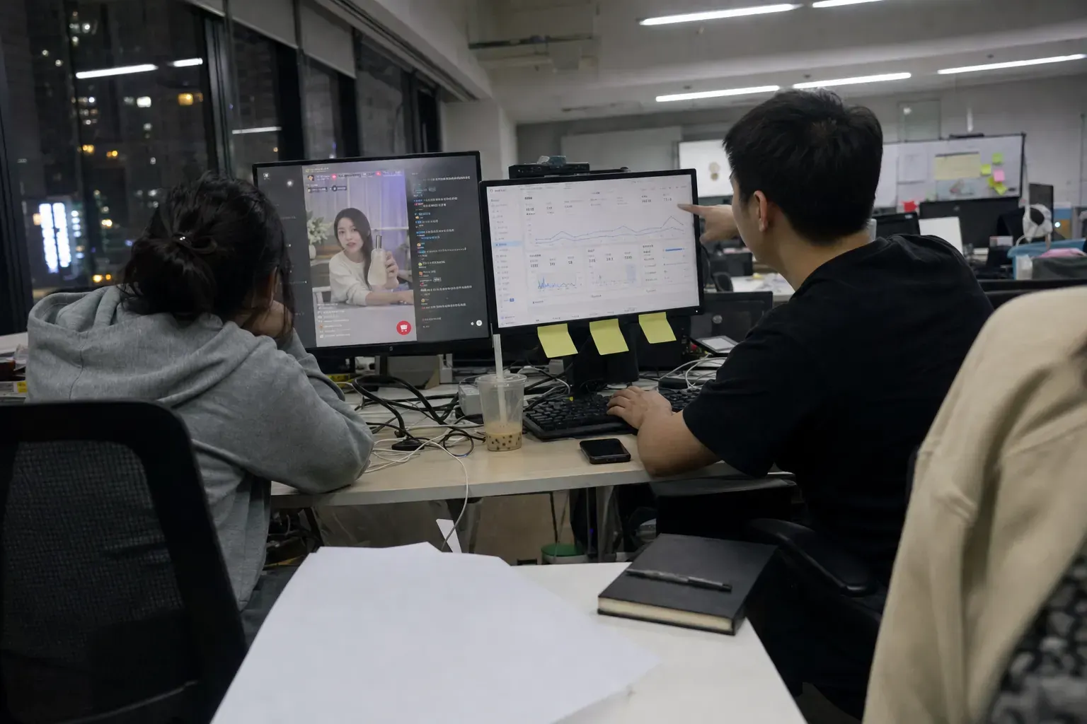 Two staff in a Chengdu Douyin agency office at night watch a brand livestream and a Douyin shop sales dashboard, blank printouts and a closed notebook on the desk