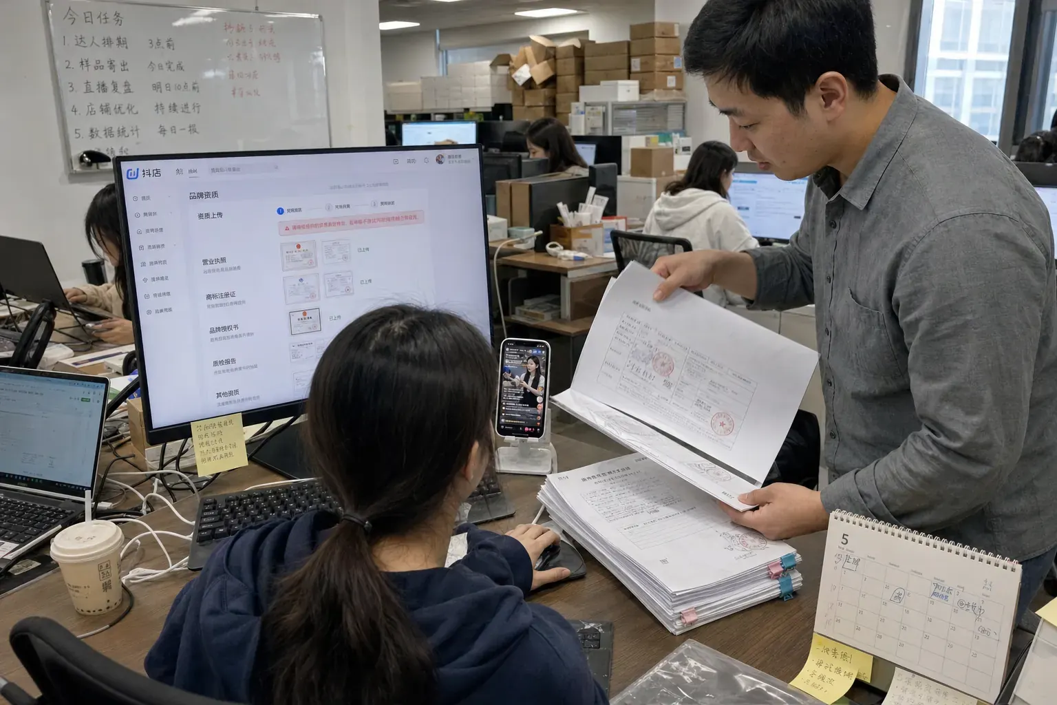 In a Hangzhou agency office, a woman uploads brand documents in the Douyin shop backend while a colleague checks a stack of stamped certificates beside her.