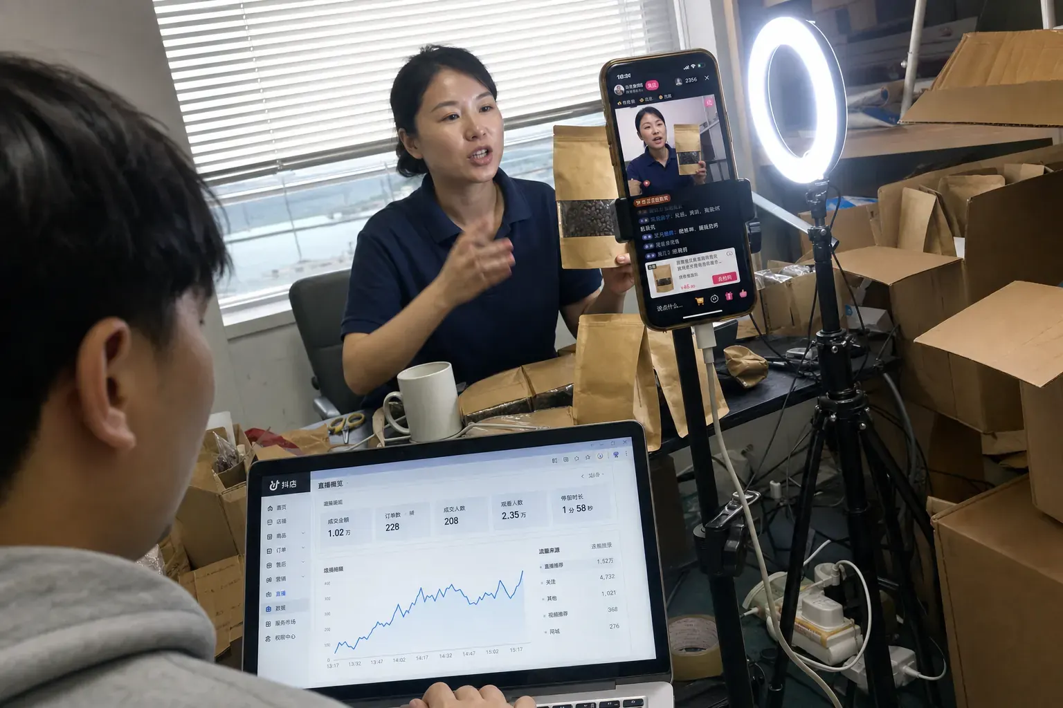 In a Xiamen brand office, a host presents coffee to a phone on a tripod in the brand's own Douyin live room while a colleague watches the shop backend on a laptop.