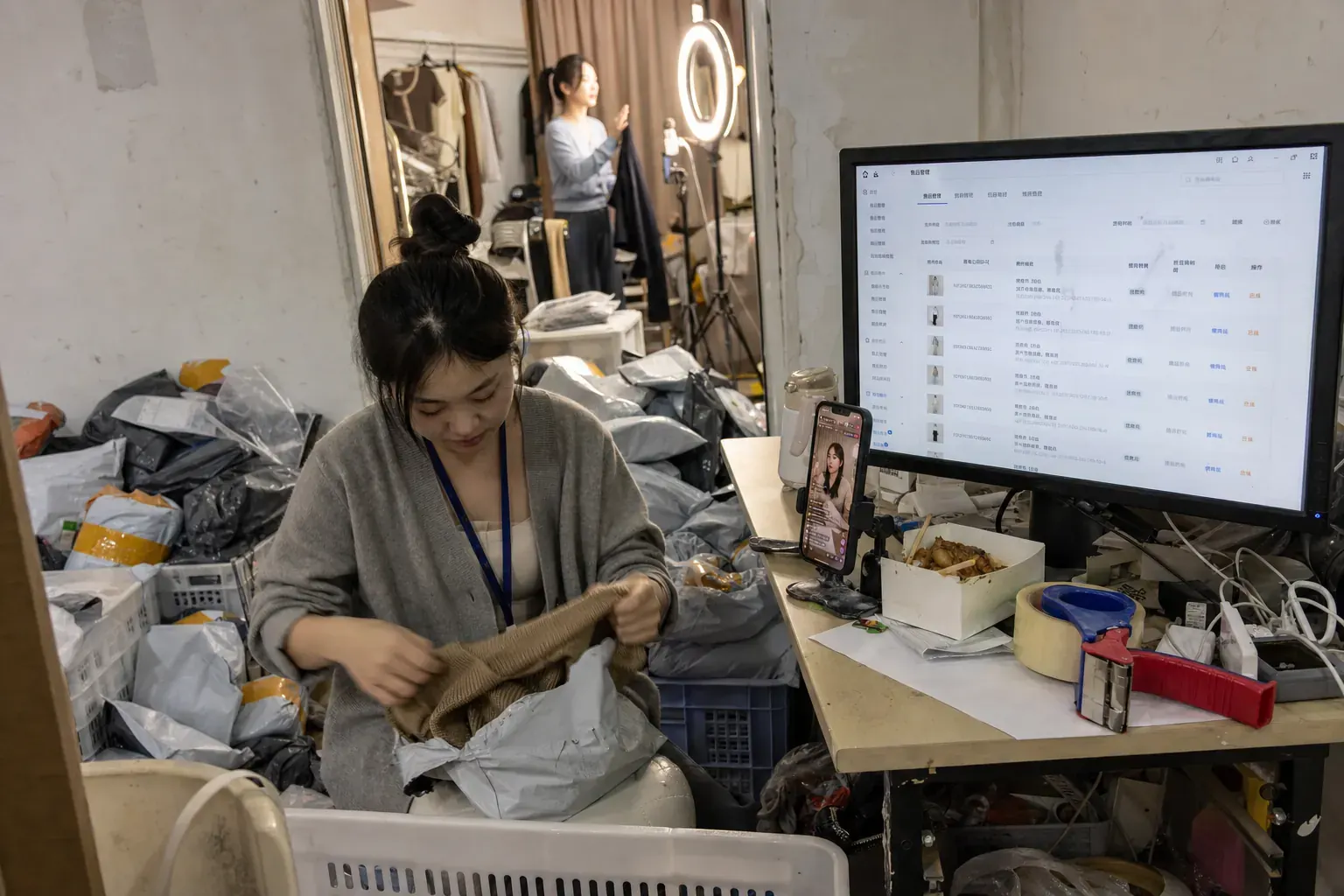 A worker in a Shenzhen clothing livestream studio unpacks a returned sweater among piles of parcels while a Douyin shop after-sale queue fills the monitor beside her