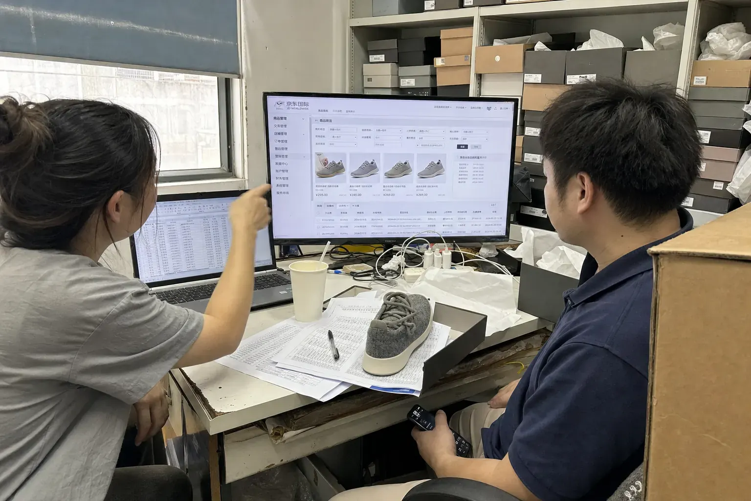 Two staff in a Guangzhou shoe stockroom look at a JD Worldwide merchant back office on a monitor, a grey wool sneaker sitting on the paperwork in front of them