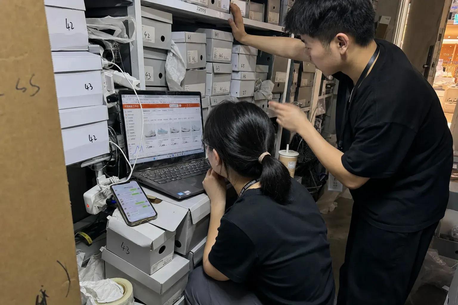 Two staff in the stockroom of a Hangzhou mall shoe shop check a Tmall seller dashboard on a laptop, surrounded by plain boxes of running shoes.
