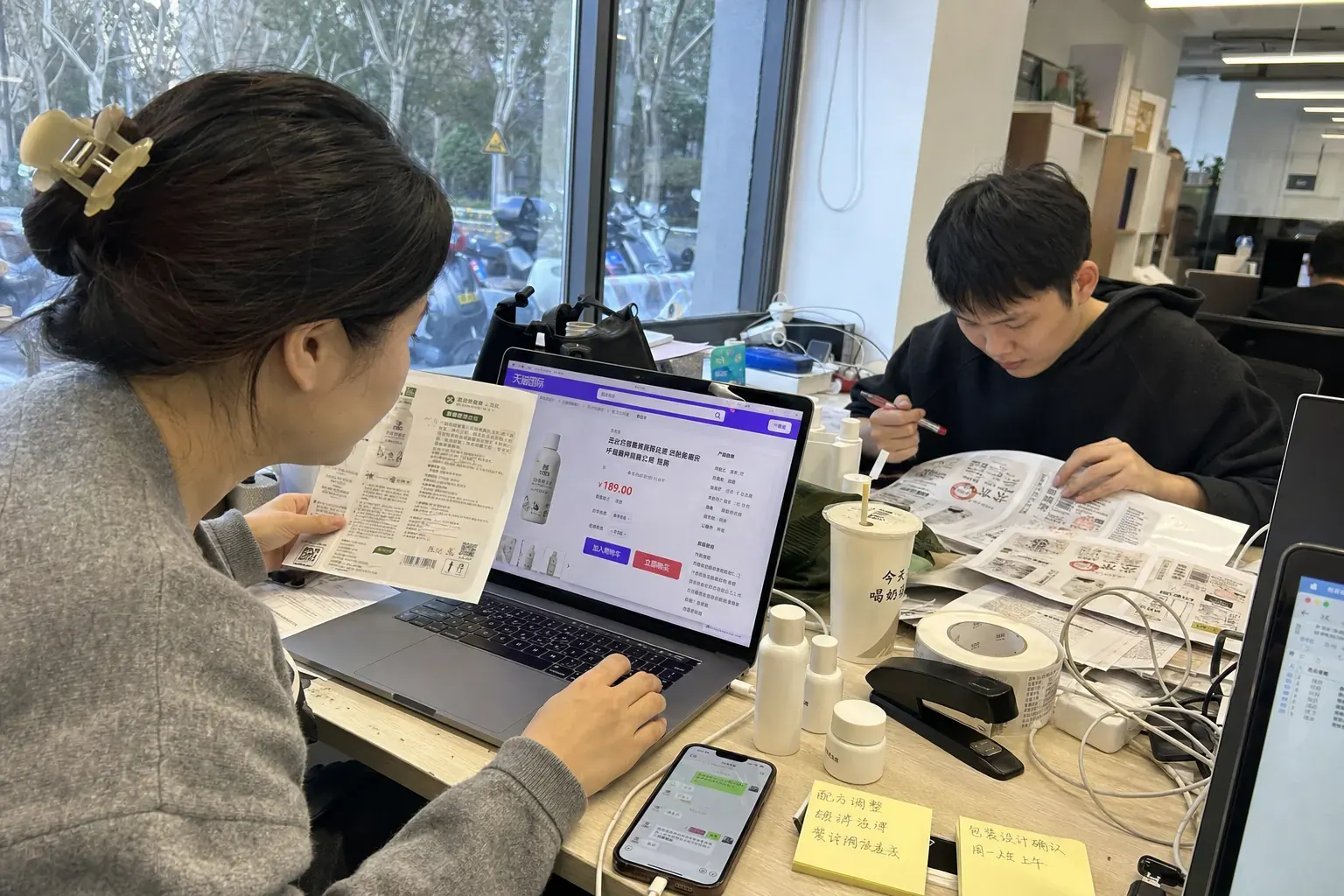 A brand manager in a Hangzhou office compares a Chinese product label with a Tmall Global product page on her laptop while a colleague marks up printed packaging proofs.