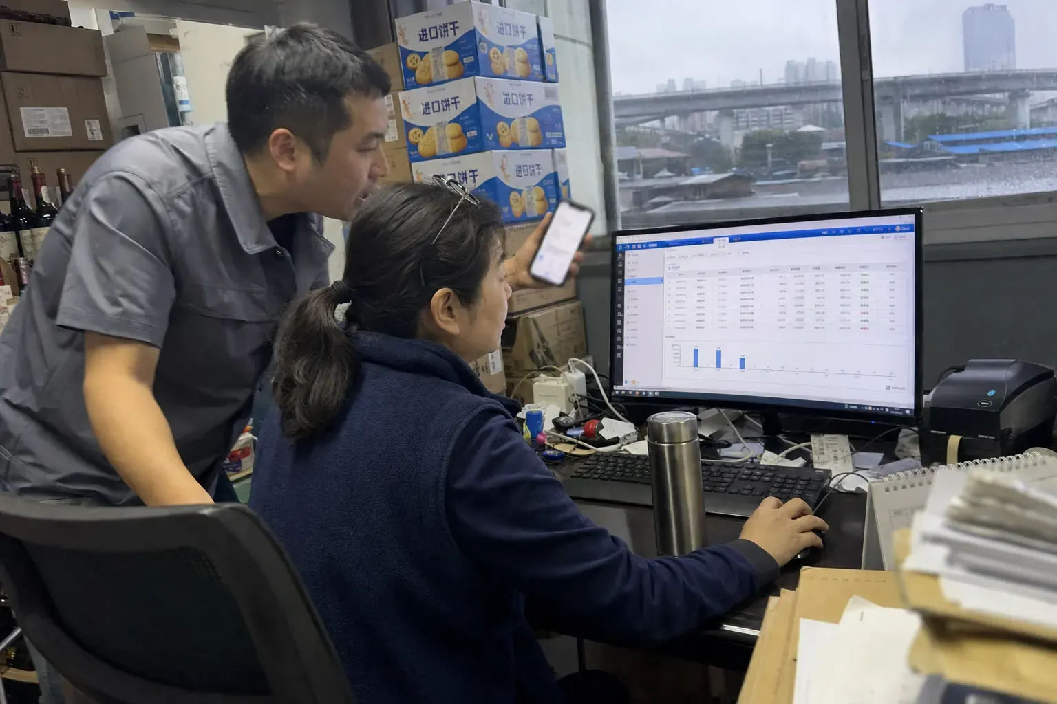 In the back office of a Wuhan food and wine importer, an accountant checks a receivables screen while a sales manager leans in holding up his phone.