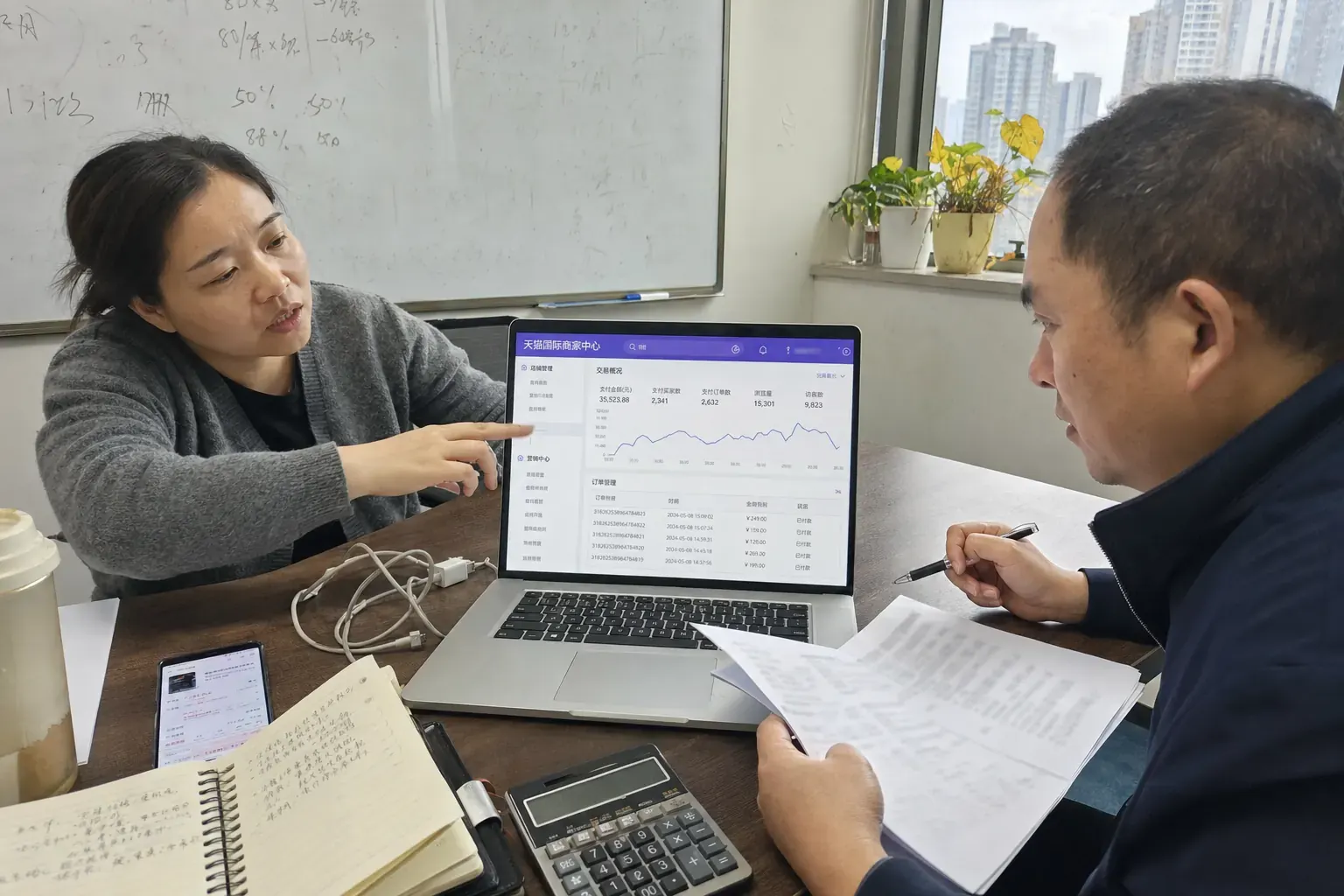 In a Changsha agency meeting room, a woman points at a Tmall Global merchant dashboard on a laptop while a man holding printed pages and a pen listens across the table.