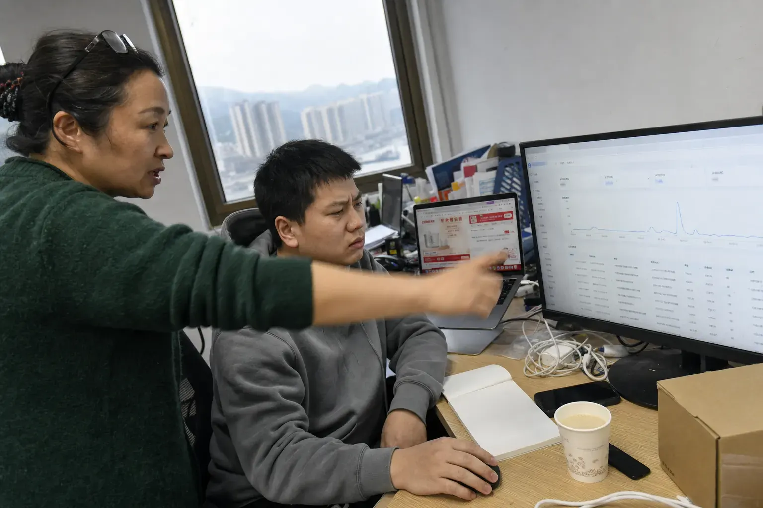In a Chongqing ecommerce office, two staff check a sudden spike in a Douyin shop's order dashboard on a desktop monitor.