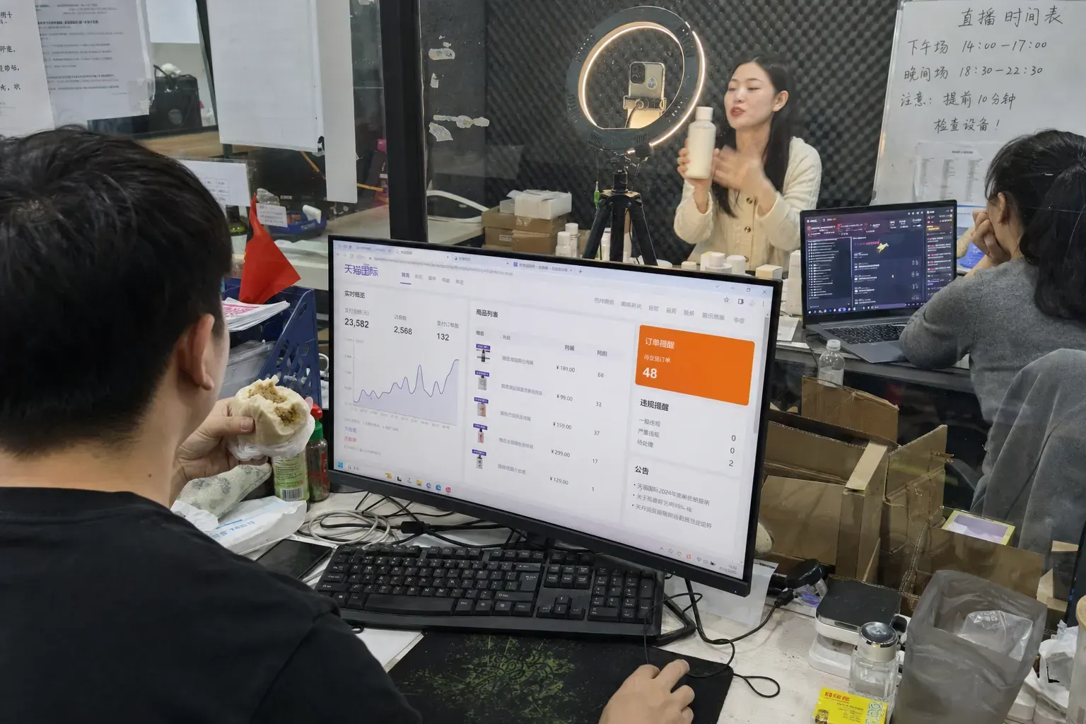 In a Guangzhou agency office, an operator eats a bun at a Tmall Global store dashboard while, behind a glass wall, a host presents a skincare bottle to a phone on a ring light for a Douyin live stream.