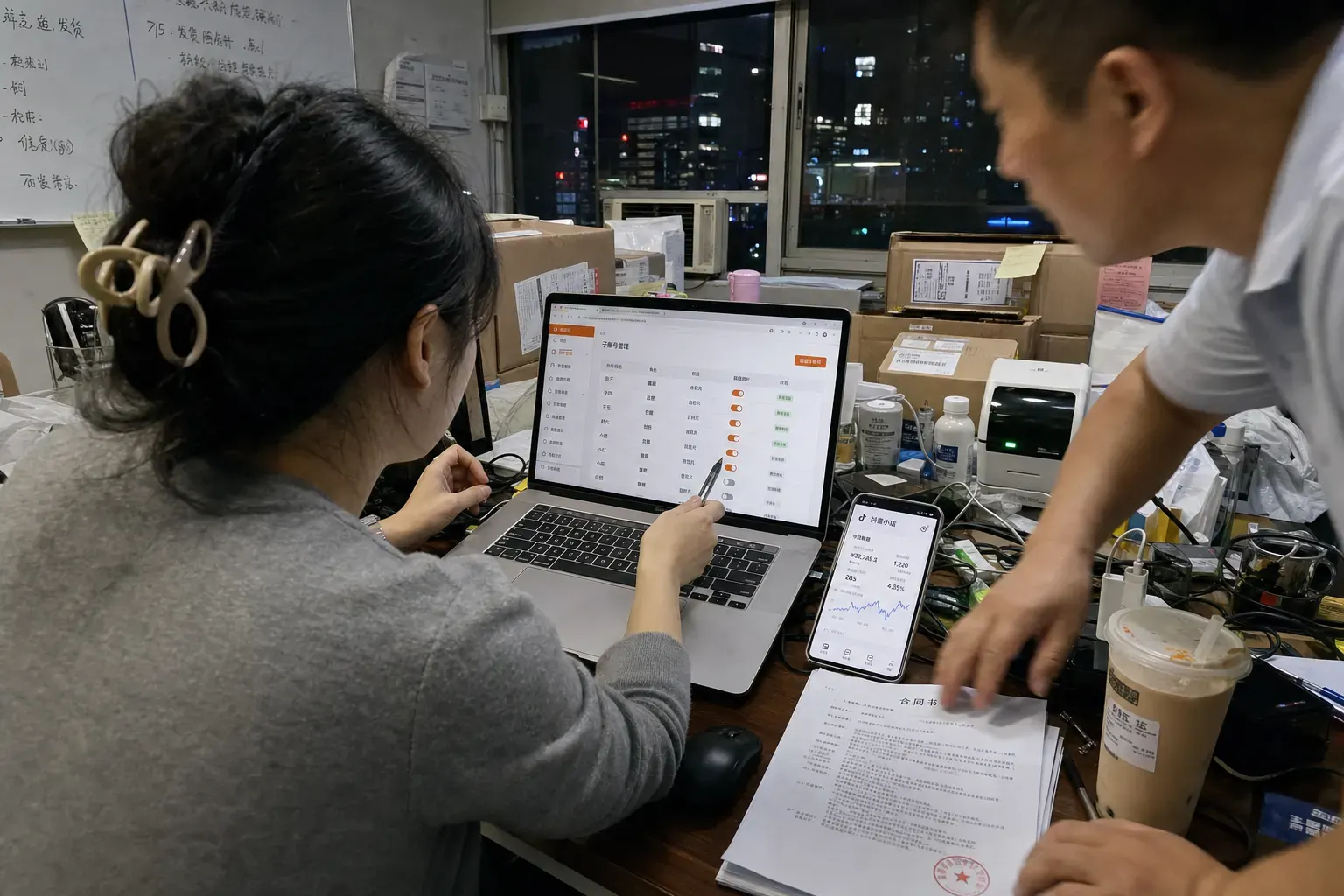 In a cluttered Shenzhen office at night, an e-commerce manager points at a sub-account management page on her laptop while a colleague reaches for a stamped contract beside a phone showing a Douyin shop dashboard.