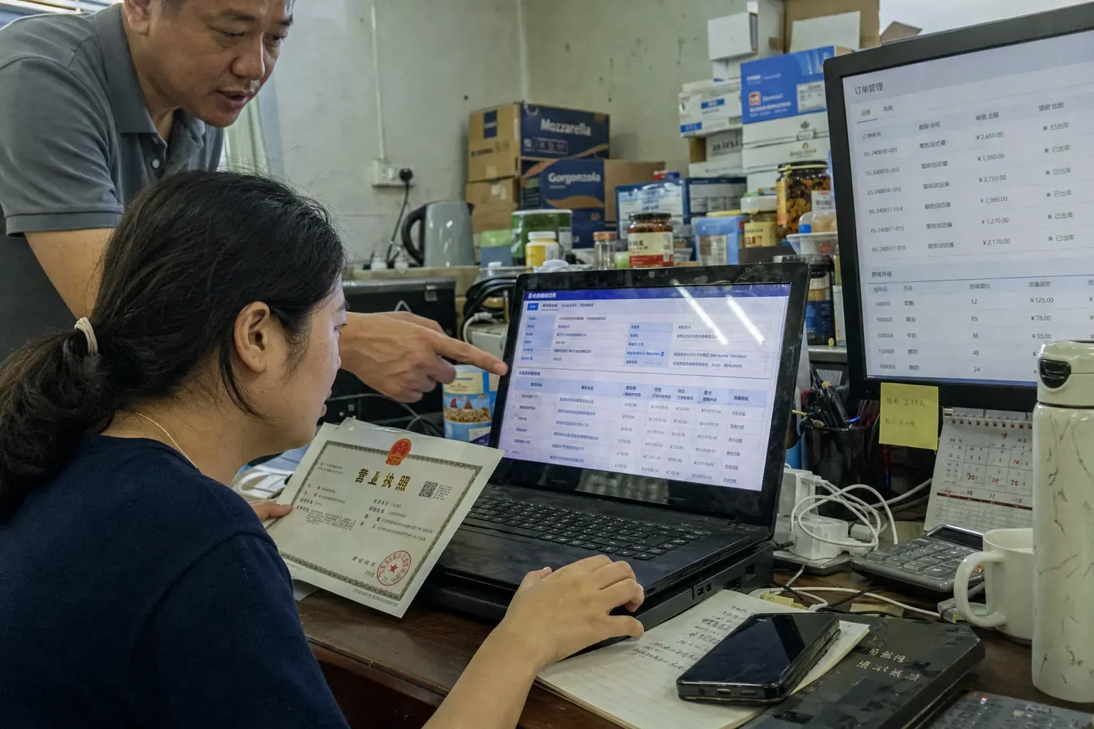 A woman at a Ningbo import distributor's office compares a Chinese company credit report on a laptop with a printed business license while a colleague points at the screen