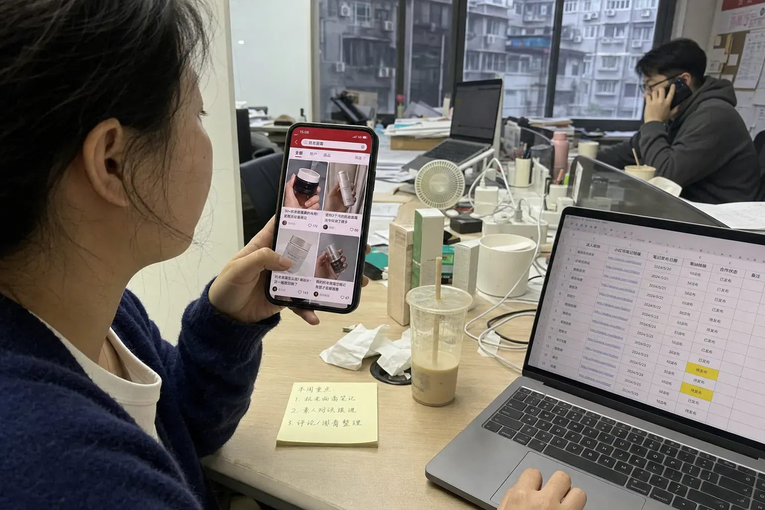 A marketer in a Chengdu office scrolls RedNote search results for a skincare product on her phone beside a laptop list of creator notes.